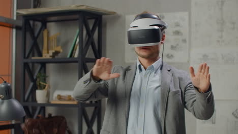 Young-man-designer-in-office-in-virtual-reality-helmet-with-hands-makes-movements-imitating-the-work-of-graphic-interface.